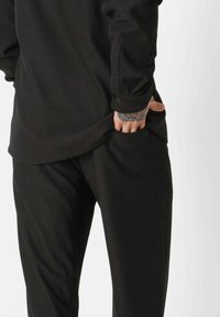 Zwarte geribbelde longsleeve shirt met knoopdetails, gecombineerd met bijpassende geribbelde zwarte broeken. De stof toont een subtiele textuur en zachtheid.