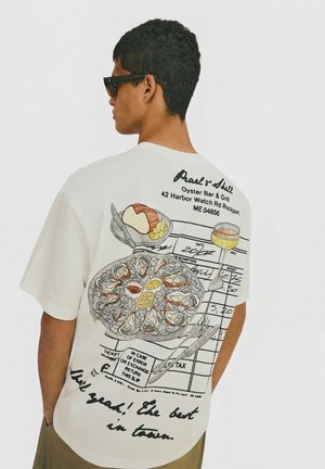 Mand iført hvid t-shirt med illustreret skaldyrsfad, brød, vinglas og restaurantkvittering trykt på ryggen.