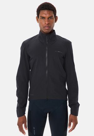 Mann trägt eine schwarze Jacke mit Reißverschluss und dunkle Sporthose, steht mit dem Gesicht nach vorne vor einem schlichten weißen Hintergrund.