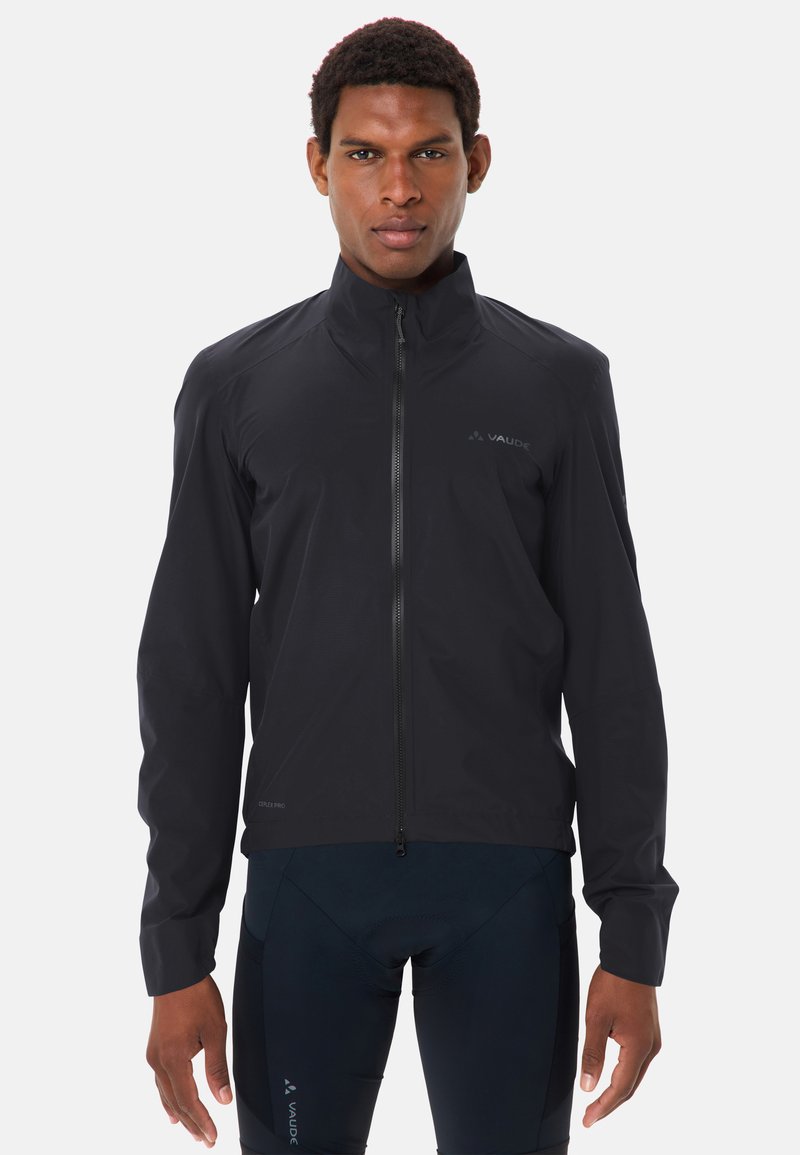 Homme portant une veste noire zippée et un pantalon de sport sombre, debout face à l'avant sur un fond blanc uni.