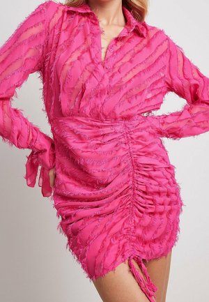 Kobieta ubrana w jaskraworóżową, teksturowaną, marszczoną mini sukienkę z długimi rękawami i kołnierzykiem, pozująca z rękami na biodrach.