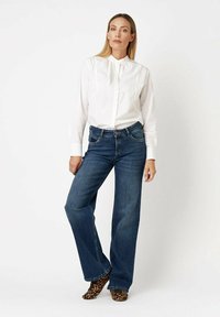 Woman wearing a white buttoned blouse, blue wide-leg jeans, and leopard print ankle boots, standing against a plain white background.