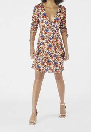 Kvinde iført en knælængde blomsterprintet slå-om-kjole med rynkede 3/4-ærmer og beige ankelstroppesandaler med høje hæle, stående mod en hvid baggrund.