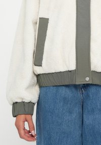 Fleece-takki, jossa on kermaista ulkomateriaalia, vihreitä yksityiskohtia ja ribbireunus. Varustettu sivutaskulla ja paikkaneulalla; pehmeä, teksturoitu pinta.