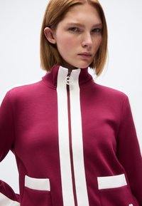 Βουργουνδίας φούτερ με φερμουάρ, λευκές λεπτομέρειες, ψηλό γιακά και δύο μπροστινές τσέπες. Ομαλή υφή· διαθέτει λογότυπο στο τράβηγμα του φερμουάρ.