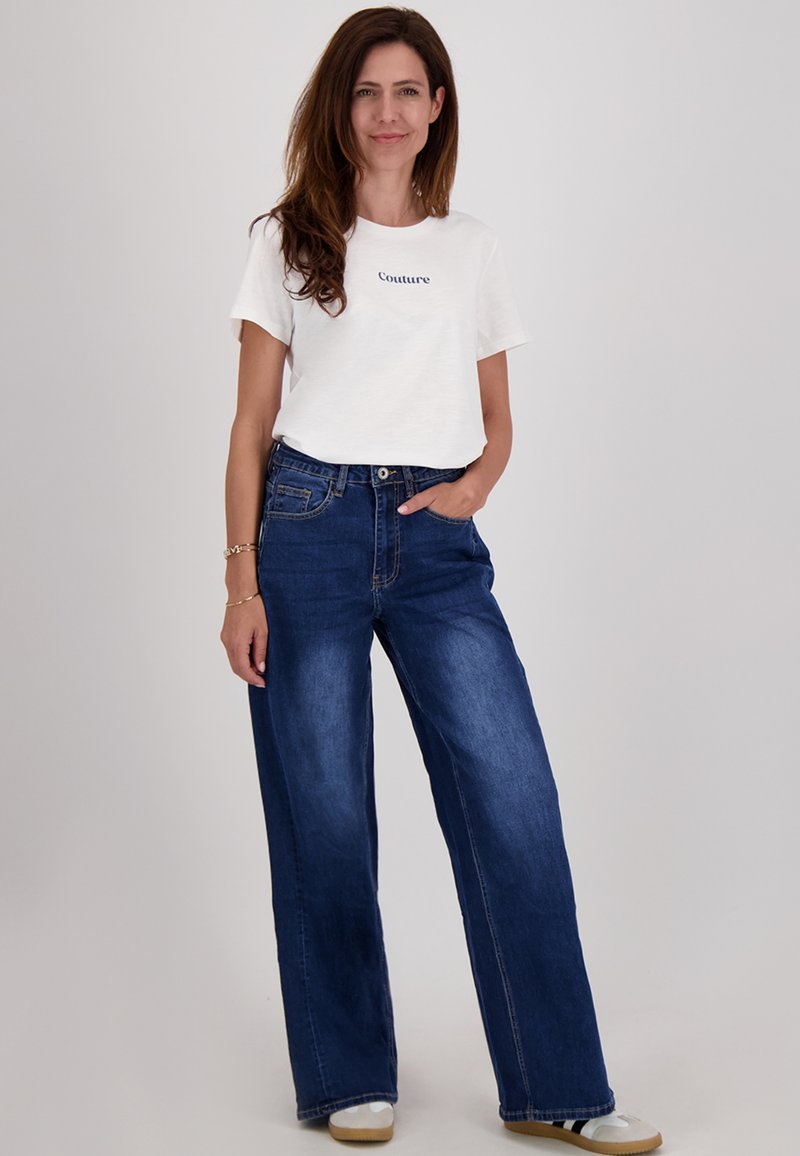 Witte t-shirt met de tekst "Couture", gecombineerd met een wijde blauwe jeans. De jeans heeft een hoge taille en subtiele vervaging, gedragen met lichte schoenen.