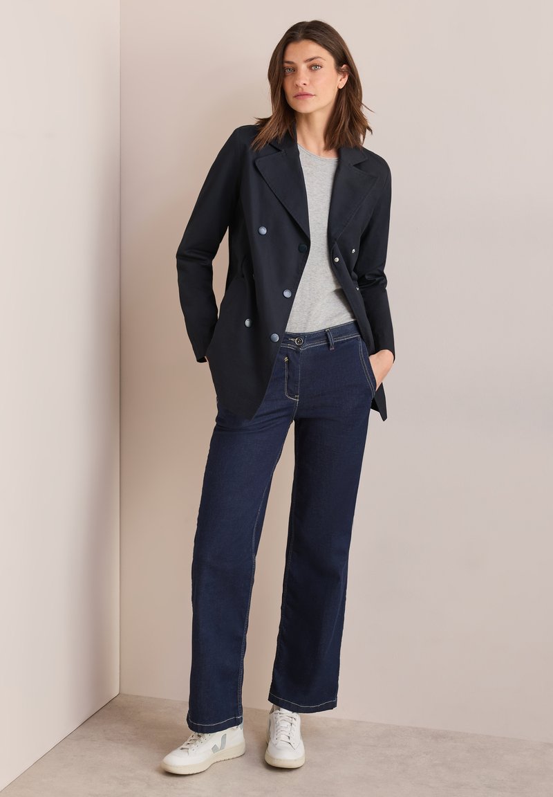 Giacca doppiopetto blu navy sopra una t-shirt grigia, abbinata a jeans larghi in denim scuro e sneakers bianche. Vengono evidenziate le trame dei tessuti lisci.