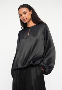 Sort satinbluse med rund halsudskæring, keyhole-detalje og ballonsleeves. Glat tekstur; kanten er let samlet ved taljen.