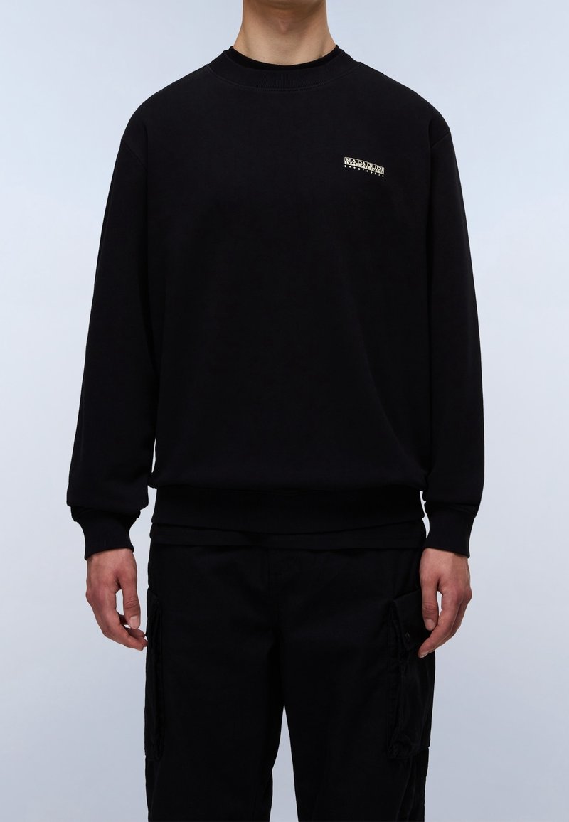 Homme portant un sweat-shirt noir à col rond avec un petit logo blanc Napapijri Geographic sur la poitrine et un pantalon cargo noir, sur un fond clair.