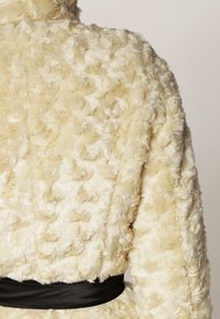 Cremfarvet, struktureret fake fur-frakke med et krøllet mønster, en sort satinbælte i taljen og en høj krave.