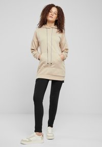 Sweat-shirt à capuche beige avec un motif texturé, doté d'une poche avant et de cordons. Associé à un pantalon noir ajusté et des baskets.
