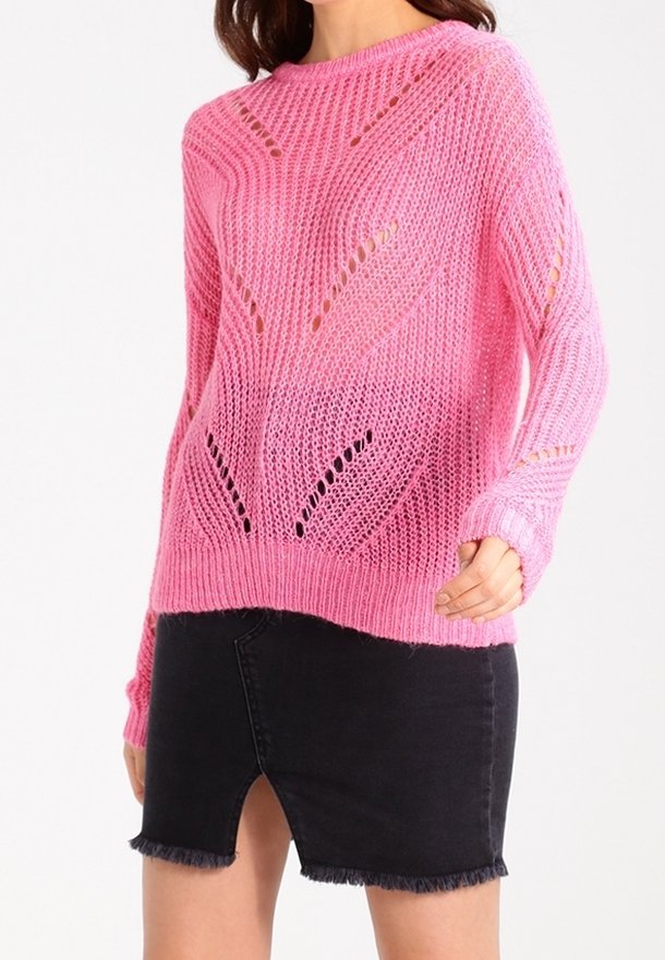 Frau in einem rosa Strickpullover mit Lochmustern und einem schwarzen Denimrock mit einem Schlitz vorne und ausgefranstem Saum.