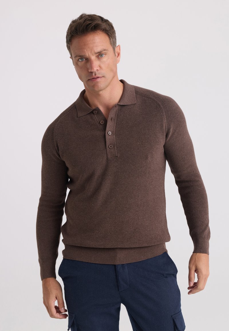 Homme portant un polo marron en maille à manches longues et un pantalon bleu foncé, debout devant un fond clair uni.
