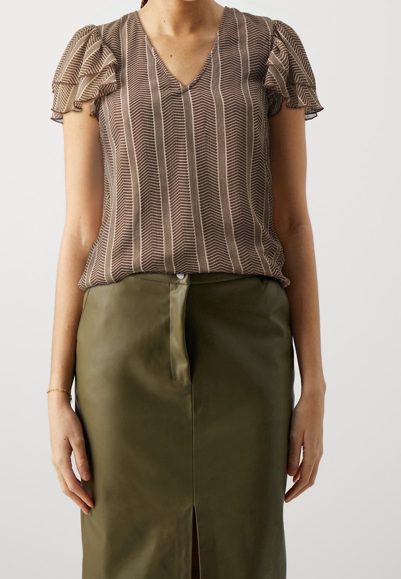 Femme portant un chemisier marron à manches courtes à motifs avec des manches bouffantes et une jupe taille haute vert olive avec une fente à l’avant.