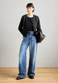 Veste noire texturée avec encolure ronde et boutons sur le devant, associée à un jean bleu clair à jambes larges. Pochette en cuir noir et chaussures.
