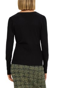 Czarny sweter z długim rękawem o dopasowanym kroju i prążkowanym dekolcie, widziany z tyłu, w połączeniu z wzorzystą zieloną spódnicą.