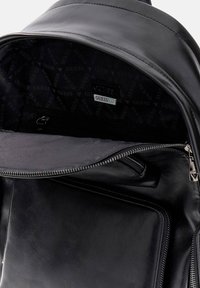 Intérieur d'un sac à dos en cuir noir avec doublure logotée, poches zippées et texture lisse. Comprend un compartiment principal et une petite poche zippée sur le côté.