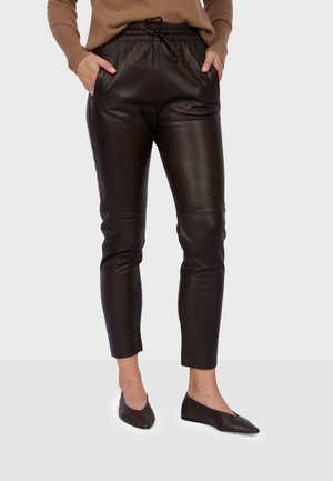 Personne portant un pantalon en cuir marron foncé avec une taille élastique et un cordon, associé à des ballerines marron assorties et un pull marron clair.
