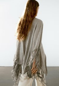 Grauer Strickponcho mit Fransen, lockerer Schnitt, V-förmiger Saum und weicher Textur, getragen über hellen Hosen.