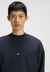 Marinblå tröja med rundad ribbad halsringning och en liten blå "HUGO"-logotyp på bröstet, tillverkad av mjukt bomullsmaterial.