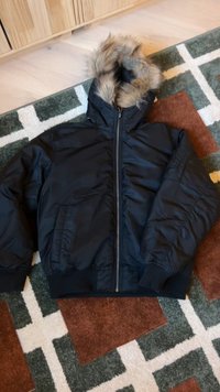 Casaco acolchoado preto com capuz forrado a pelo, fecho frontal com fecho éclair e punhos canelados. Apresenta bolsos laterais e uma textura lisa e mate.