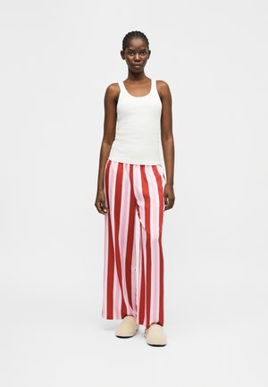 Femeie stând purtând un maiou alb, pantaloni largi cu dungi roșii și roz și papuci bej, pe un fundal simplu și luminos.