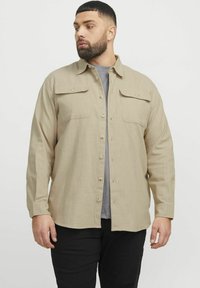 Beige overhemd met knopen, voorzien van twee borstzakken, lange mouwen en een rechte zoom. Draag het over een grijskleurig t-shirt met een gestructureerde stof.