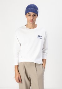Hvit langermet skjorte med blå logo, kombinert med beige plisserte bukser. En blå ribbestrikket lue fullfører antrekket.