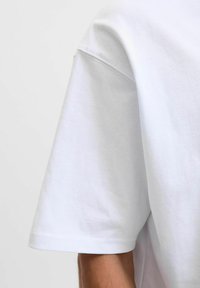 Prim-plan cu o persoană care poartă un tricou simplu alb cu mânecă scurtă, arătând cusătura de la mânecă și umăr.
