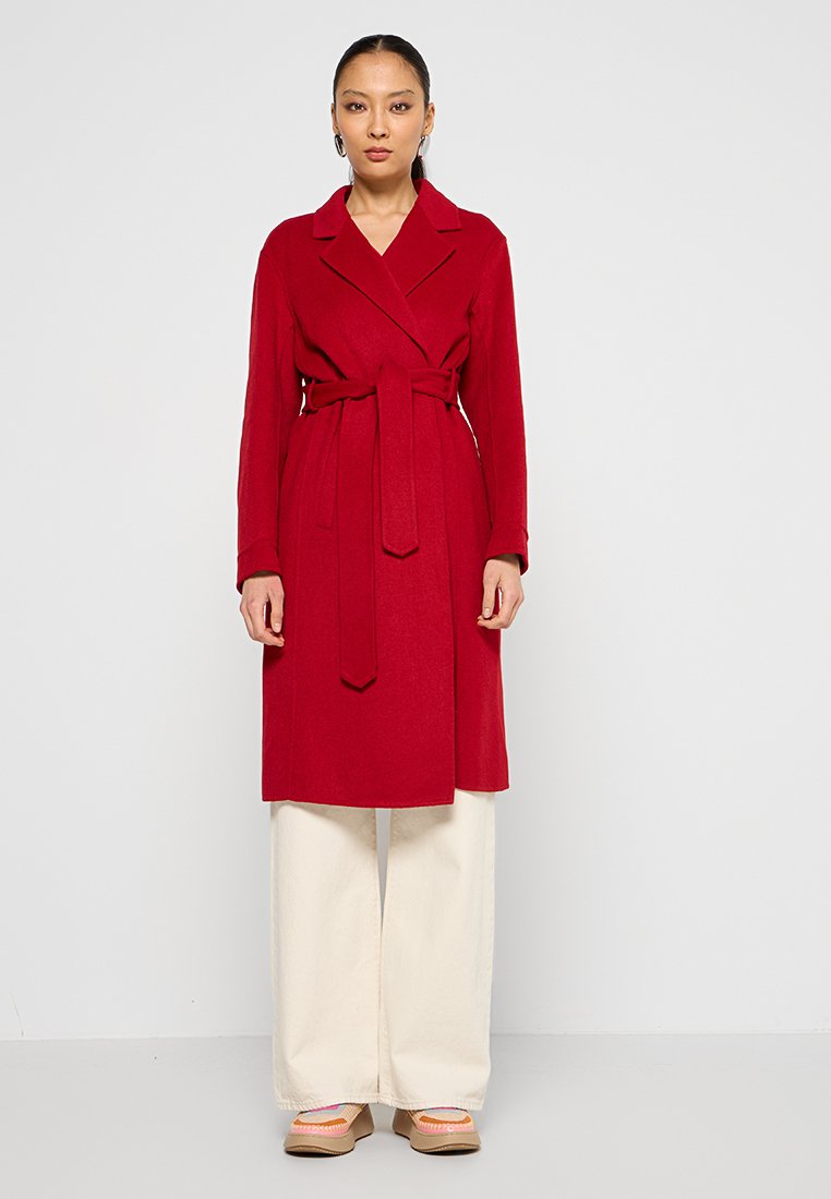 Roter Woll-Trenchcoat mit Gürtel, klassischem Kragen und langen Ärmeln. Kombiniert mit weiten weißen Hosen und beigen Slippers.