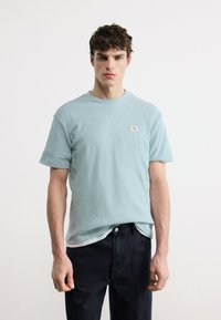 Νέος άνδρας με κοντά σγουρά μαλλιά, που φοράει ένα γαλάζιο υφασμάτινο μπλουζάκι και μαύρο τζιν, στέκεται μπροστά σε ένα απλό λευκό φόντο.