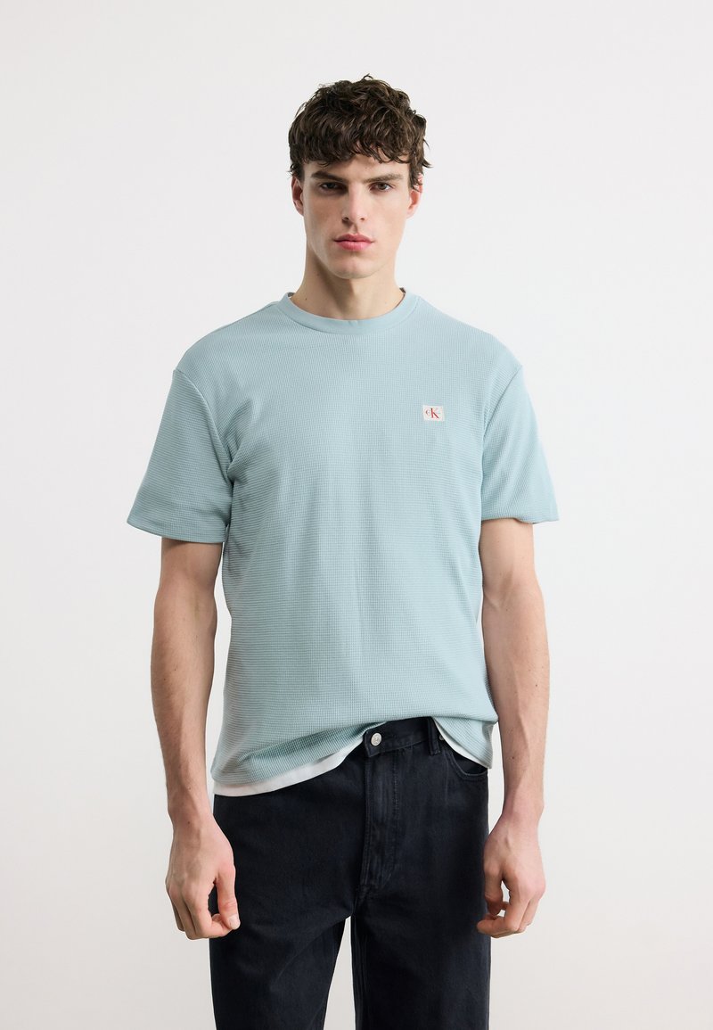 Νέος άνδρας με κοντά σγουρά μαλλιά, που φοράει ένα γαλάζιο υφασμάτινο μπλουζάκι και μαύρο τζιν, στέκεται μπροστά σε ένα απλό λευκό φόντο.