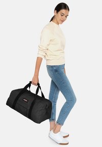 Musta duffel-laukku, jossa on kaksoiskahvat, valmistettu kestävästä kankaasta. Sisältää vetoketjullisen taskun ja logopatchin. Malli pitää laukkua rentoissa vaatteissa.