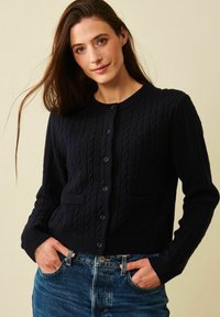 Marinblå stickad cardigan med kabelmönster, ribbade ärmslut och nederkant, rund hals och sex framknappar. Har två framfickor.