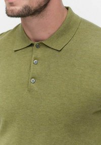 Oliivinvihreä polo-paita, joka on valmistettu teksturoidusta kankaasta. Paidassa on kaulus ja kolme painonappia, joissa on pyöreät harmaat napit.
