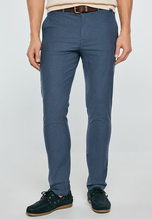 Mężczyzna w niebieskich, dopasowanych spodniach z brązowym paskiem, beżowym swetrze i ciemnoniebieskich, casualowych butach stojący na jasnym tle.