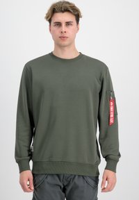 Sudadera verde oliva con mangas largas, cuello redondo acanalado y un bolsillo con cremallera en el brazo izquierdo que presenta un detalle de etiqueta roja.