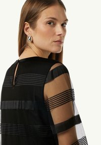 Schwarzes Kleid mit hochgeschlossenem Ausschnitt, transparenten gestreiften Ärmeln und einem Schlüsselspalt im Rücken mit einem Knopfdetail. Der Stoff hat eine glatte Textur mit einem geschichteten Look.