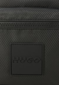 Black textured bag with a zipper and a square rubber logo patch embossed with the word "HUGO" in the center.
