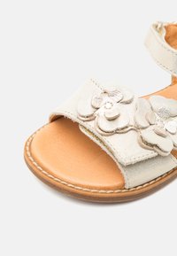 Sandal alb din piele cu ornamente florale, talpă interioară de nuanță deschisă, cu detalii cusute pe talpă. Curea ajustabilă pentru o adaptare perfectă.