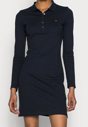 Femme portant une robe polo bleu marine à manches longues avec un petit logo sur la poitrine gauche et des boutons au col.