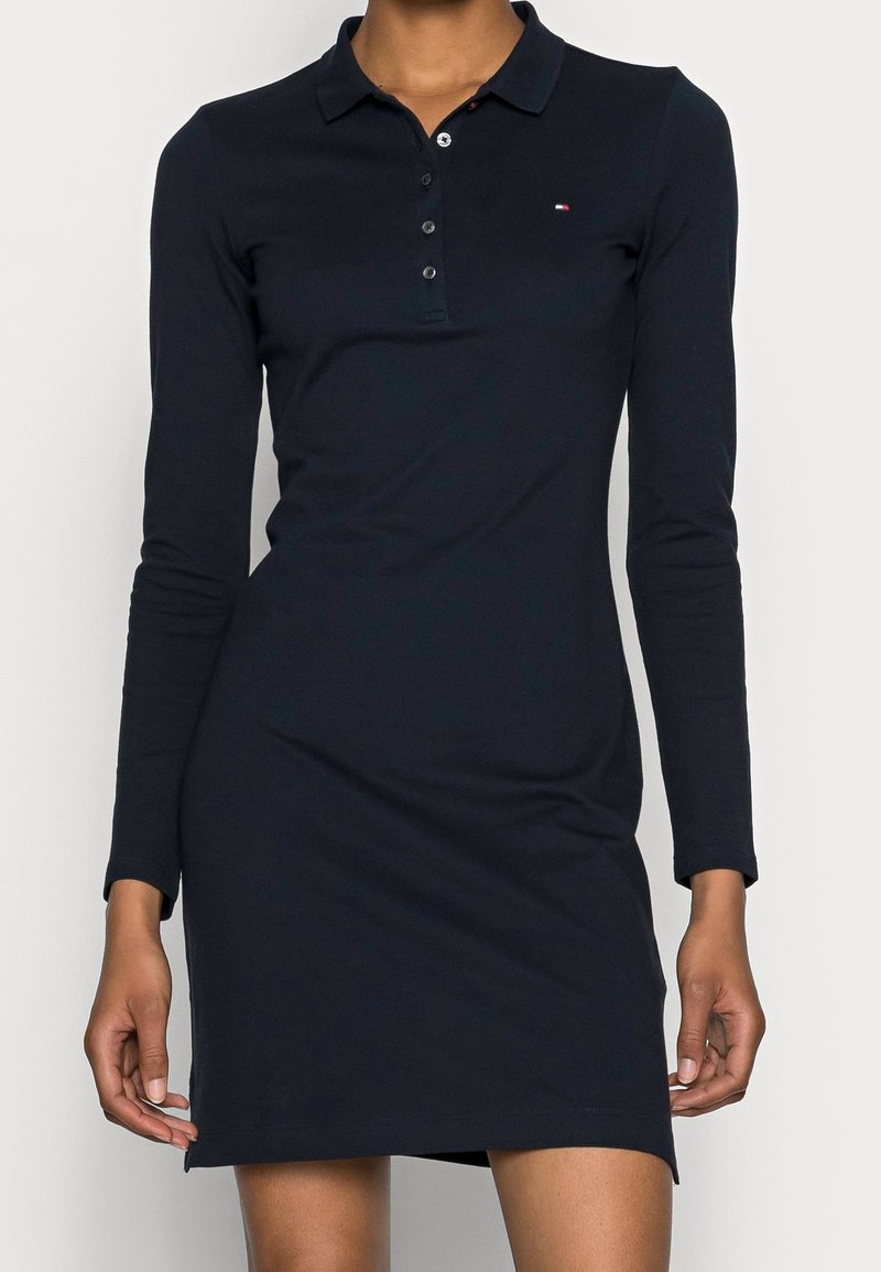 Femme portant une robe polo bleu marine à manches longues avec un petit logo sur la poitrine gauche et des boutons au col.