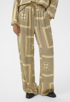 Bézs színű bőszárú nadrág fekete geometrikus négyzet és kör mintával, elasztikus derékrésszel, húzózsinórral, fekete cipővel viselve.