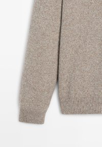Beige stickad tröja med en mjuk textur, med ribbad manschett och fåll. Tyget har en subtil melerad blandning för visuell intresse.