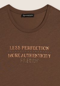 T-shirt in cotone marrone con scollatura rotonda, con la scritta "MENO PERFEZIONE PIÙ AUTENTICITÀ" in carattere metallico e strass decorativi.