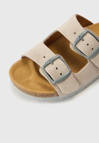 Világos bézs színű, velúr szandál két állítható pánttal, szürke csatokkal és barna texturált talpbéléssel. Lapos talp szürke külső peremmel.