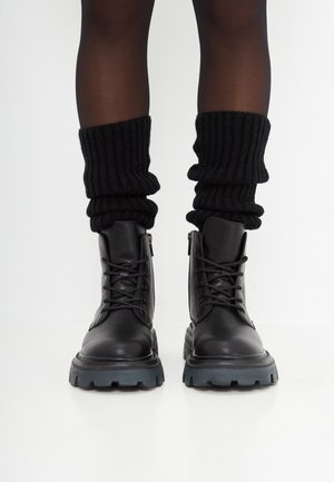 Botas negras de cuero con cordones y suelas gruesas, usadas con calentadores de piernas de punto acanalado negros sobre medias oscuras en una persona de pie.