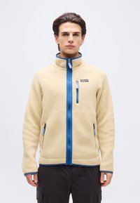 Νεαρός άνδρας που φοράει μπεζ φλις μπουφάν με μπλε φερμουάρ και λεπτομέρειες, στέκεται μπροστά σε απλό φωτεινό φόντο, με τα χέρια χαλαρά στα πλευρά.