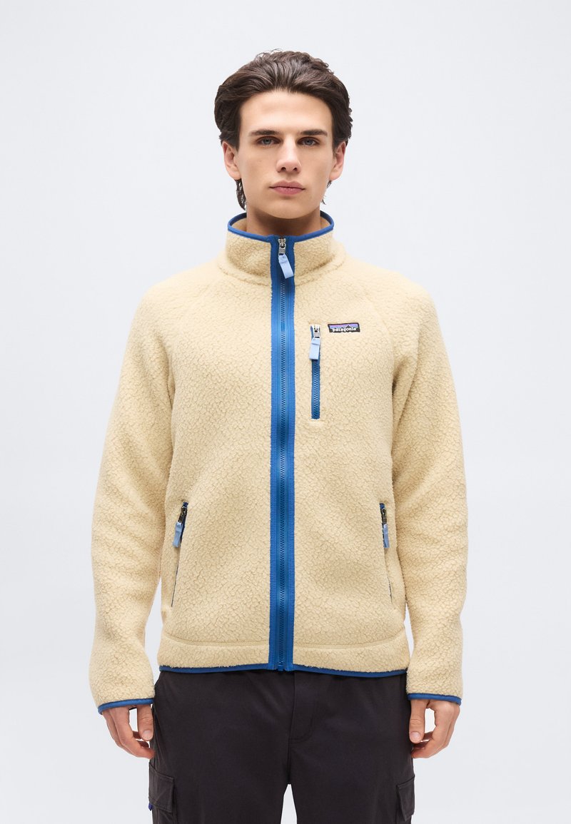 Νεαρός άνδρας που φοράει μπεζ φλις μπουφάν με μπλε φερμουάρ και λεπτομέρειες, στέκεται μπροστά σε απλό φωτεινό φόντο, με τα χέρια χαλαρά στα πλευρά.