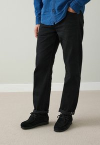 Svarta jeansbyxor i rak passform med uppvikta muddar, kombinerade med svarta mockaskor, under en blå strukturerad skjorta.